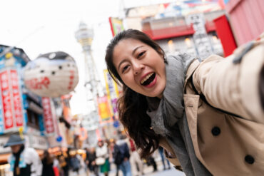 female taking picture with tsutenkaku tower self portrait of happy Asian Japanese female tourist looking at camera while taking picture with tsutenkaku tower at background in nostalgic Shinsekai shopping area of Osaka Japan