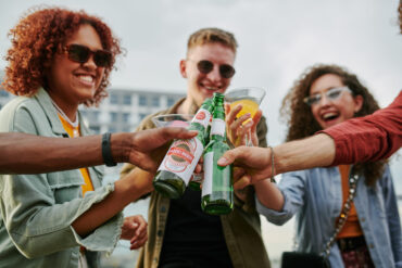 Group of young happy multicultural people clinking with beer and cocktails Group of young happy multicultural people clinking with bottles of beer and cocktails while enjoying their gathering in rooftop cafe